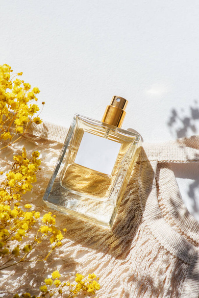 Διαφανές μπουκάλι άρωμα με ετικέτα και υφασμάτινη τσάντα για ψώνια σε λευκό φόντο. Παρουσίαση αρώματος με το φως της ημέρας. Μοντέρνο σχέδιο σε φυσικά υλικά με κίτρινα άνθη αγρού. Η ουσία των γυναικών και των ανδρών. - Φωτογραφία, εικόνα