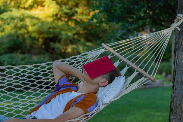 Jovem descansando e dormindo em uma rede na primavera com um livro vermelho cobrindo seus olhos e rosto no jardim - Foto, Imagem