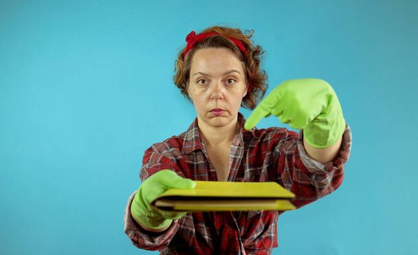 Una mujer de cuarenta años con guantes limpiadores de goma señala con el dedo un libro. Una mujer adulta sobre un fondo azul sostiene un libro amarillo. Retrato de mujer pin-up - Foto, Imagen