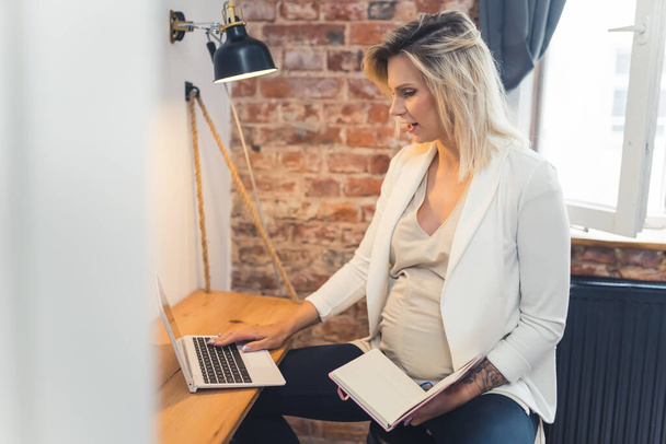 Pregnant but still active at work. Middle-aged blond Caucasian woman in a white blazer sitting at her office desk, working on a laptop, and holding a paperback calendar. High quality photo - Foto, Imagen