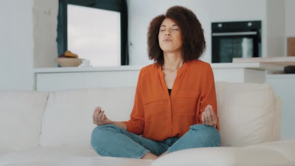 Afrikkalainen amerikkalainen rauhallinen nainen istuu lootuksen asennossa mukavalla sohvalla olohuoneessa kotona suljetuin silmin kihara tukka tyttö meditaatio käytäntö harmonia henkinen tasapaino meditointi mindfulness ei stressiä - Materiaali, video