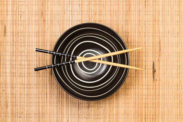 Chopsticks on a black bowl and on a bamboo table mat in a top view - Foto, imagen