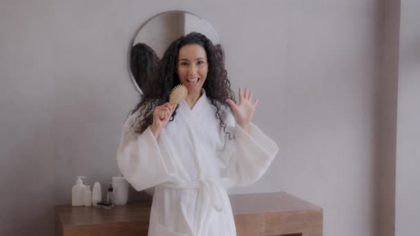 Young happy carefree woman in bathrobe dancing in bathroom expressive sings using hairbrush as microphone active funny hispanic girl moving dynamic shaking chic long curly hair enjoying daily haircare - 映像、動画