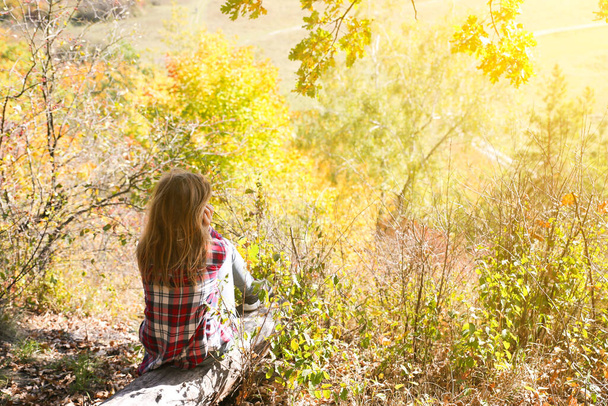 Girl in the checkered shirt is sitting in the autumn forest. Seasonal concept. Stylish hipster clothes outdoors. Nature philosophy around. Beautiful woman near the yellow leaves and trees. - Photo, image