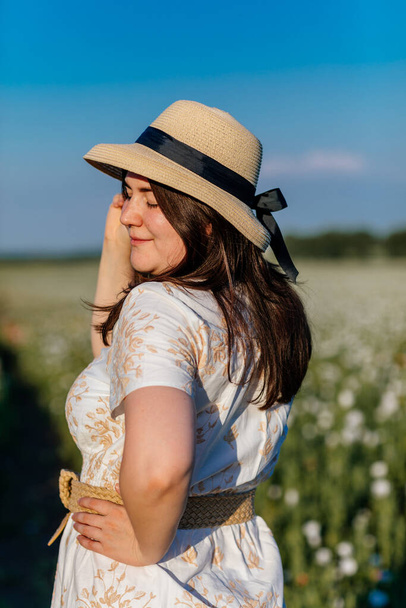 Scenic picturesque of beautiful girl in white and beige dress and straw hat in poppy field at sunset, Portrait of romantic young woman with flowers, body positivity, Selective focus natural light - Photo, image