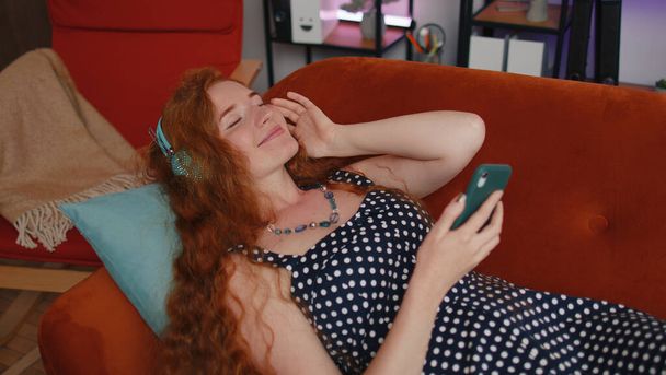 Happy overjoyed redhead young girl in wireless headphones relaxing lying on sofa at home choosing listening favorite energetic disco rock n roll dancing music in smartphone. People weekends activities - Fotó, kép