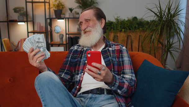 Planowanie budżetu rodzinnego. Uśmiechnięty dziadek liczący pieniądze w gotówce, używający smartfona do obliczenia rachunków krajowych w domu. Radosny człowiek zadowolony z dochodów i oszczędza pieniądze na planowane wakacje, prezenty - Zdjęcie, obraz