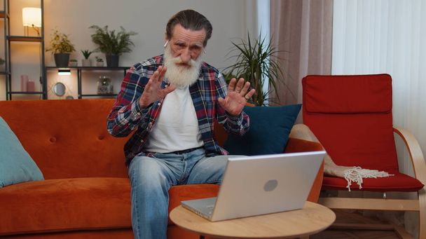 Der ältere Großvater sitzt auf der Couch, schaut auf den Laptop, macht Videokonferenzen mit Freunden oder der Familie und genießt angenehme Gespräche. Älterer Mann lacht und winkt allein zu Hause Hallo - Foto, Bild