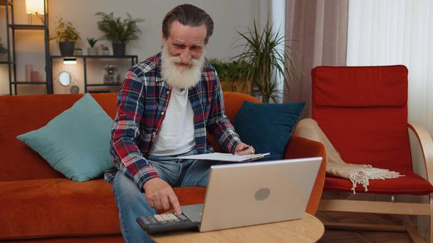 Glücklich lächelnder älterer Großvater, der Zahlungen abrechnet und monatliche Spesen berechnet, den Papierkram auf der heimischen Couch analysiert. Ältere Freiberufler arbeiten online Remote-Job auf Laptop - Foto, Bild
