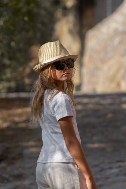 A beautiful young girl with blond hair in a hat and sunglasses turned around and looked at the camera. - Foto, afbeelding