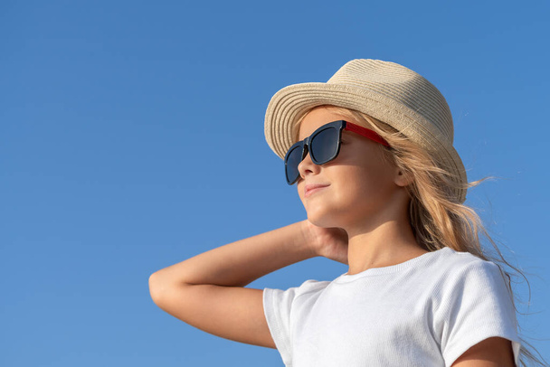 A young girl in a hat and sunglasses straightens her hair with one hand against the blue sky and looks to the side. - Photo, image