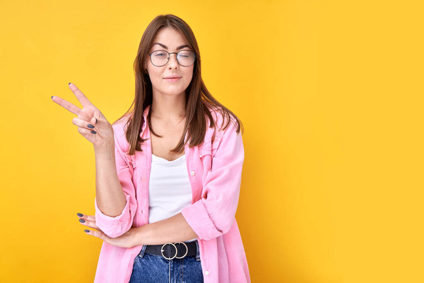 Portrait of positive brunette girl showing peace gesture and winking isolated on yellow background - Foto, Imagen