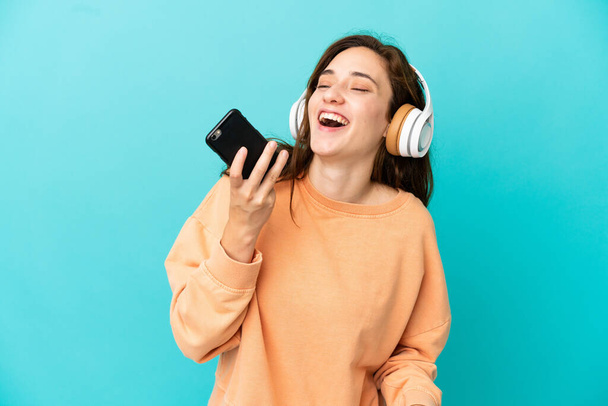 Young caucasian woman isolated on blue background listening music with a mobile and singing - 写真・画像
