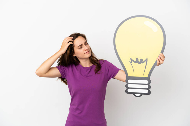 Young caucasian woman isolated on white background holding a bulb icon and having doubts - Φωτογραφία, εικόνα