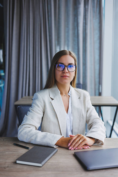 Успешная молодая женщина-менеджер в элегантной повседневной куртке и оптических очках, сидит в офисе на рабочем месте, опытная женщина с современным ноутбуком, работающая в офисе - Фото, изображение