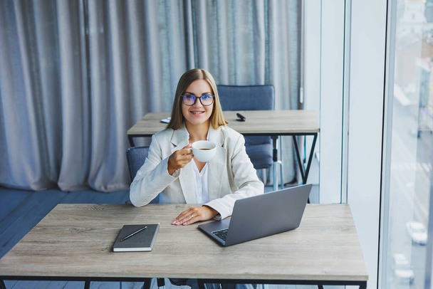 Χαρούμενη μάνατζερ γυναίκα σε γυαλιά με φορητό υπολογιστή χαμογελώντας στην κάμερα κατά τη διάρκεια του διαλείμματος καφέ στο εσωτερικό του γραφείου, χαρούμενη Ευρωπαία γυναίκα με ποτό καφεΐνης ποζάροντας - Φωτογραφία, εικόνα