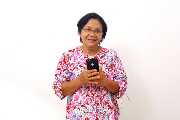 Asiatische ältere Frau im Stehen, während sie ein Handy in der Hand hält. Isoliert auf weißem Hintergrund - Foto, Bild