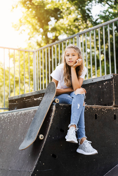 Child girl sitting with skate board on sport ramp. Sports equipment for kids. Active teenager with skate board on skate park playground - Foto, afbeelding