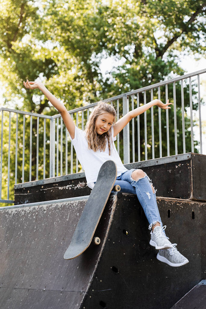Having fun with skate board. Funny child girl with skate sitting on sport ramp, smiling and grimacing on skate playground. Active teenager posing with skate board. Extreme lifestyle - Zdjęcie, obraz