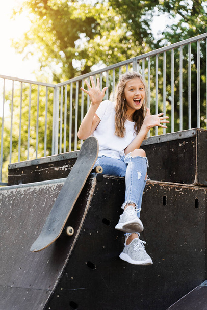 Having fun with skate board. Funny child girl with skate sitting on sport ramp, smiling and grimacing on skate playground. Active teenager posing with skate board. Extreme lifestyle - 写真・画像