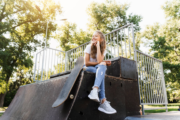 Child girl sitting with skate board on sport ramp. Sports equipment for kids. Active teenager with skate board on skate park playground - Fotografie, Obrázek