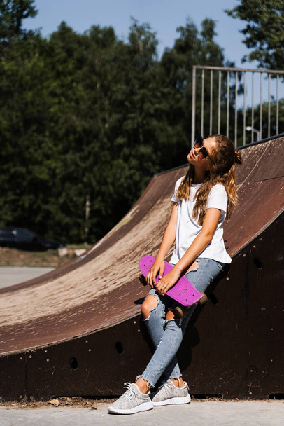Dziewczyna moda dziecko w okularach pozowanie z tablicy groszowej na skate placu zabaw. Aktywny nastolatek ze sprzętem sportowym na rampie sportowej. Ekstremalny styl życia - Zdjęcie, obraz
