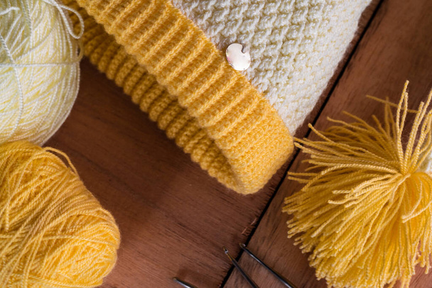 yellow and white cap for winter, hand knitted in crochet on an orange wooden background, next to it two rolls of wool and three steel needles ready to work at home. concept of enterprise - Zdjęcie, obraz