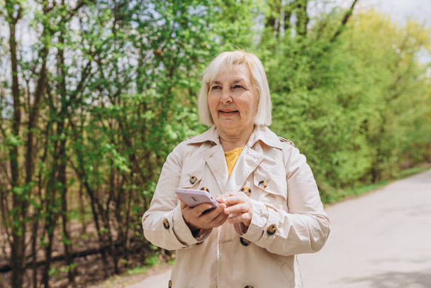 Ώριμη ξανθιά γυναίκα σε casual ρούχα περιήγηση έξυπνο τηλέφωνο, ενώ πάρκο της πόλης. Καυκάσιος blogger κρατά το τηλέφωνο σκεπτόμενος την ιδέα για κοινωνική δημοσίευση σε εξωτερικούς χώρους - Φωτογραφία, εικόνα