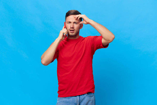 赤いTシャツの混乱した日焼けしたハンサムな男は,ブルースタジオの背景に隔離された額に触れる債務収集者からの電話で悪いニュースを得ます. スペースモックアップをコピー. オンラインライフスタイルコンセプト - 写真・画像