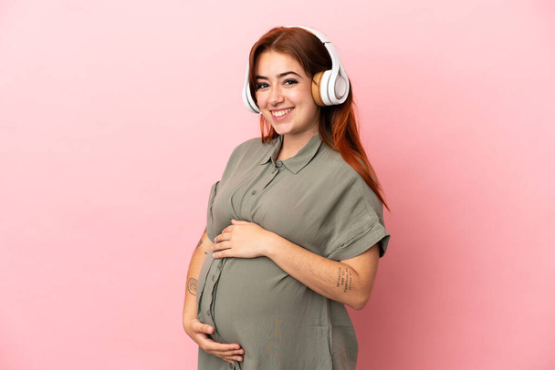 Young redhead caucasian woman isolated on pink background pregnant and listening music - Foto, afbeelding