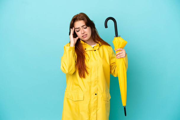 Redhead woman holding an umbrella isolated on blue background with headache - Foto, Bild