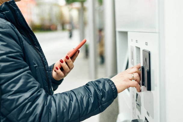 Woman buying ticket at ticket machine paying using mobile payment app on smartphone. Female buying bus public transpoort ticket in vending machine. Person purchasing car parking ticket using mobile phone - Zdjęcie, obraz