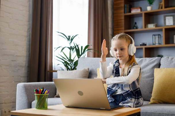 Primary schoolgirl wearing earphones has online lesson, video call with teacher, raising hand to answer the question. Cute child studying at home using laptop. Distance learning. Home education. - Fotó, kép