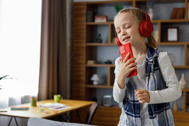 Happy little girl wearing red earphones dancing and singing using smartphone like microphone. Cute child having fun jumping on coach. Home leisure, happy kid. Attention deficit hyperactivity disorder - Photo, Image