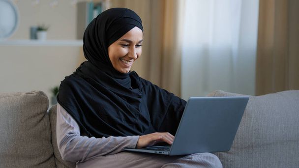 ハッピーアラブ女性イスラム教徒の若い女の子の女性ヒジャーブでフリーランスのクリエイターは、自宅でコンピュータのテキストメッセージの電子メールの仕事で友人とチャットに座るインターネットでのオンライン検索求人空室Webデザインの電子商取引を開発 - 写真・画像