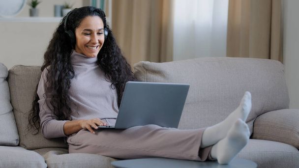 Hiszpanka arabski słodkie kobieta dziewczyna nosić słuchawki siedzieć na kanapie w domu pokój dzienny trzymać laptop używać rozmowy wideo odległość fala pozdrowienia chłopak uśmiechnięty sieci społecznej komunikacji podczas pandemii - Zdjęcie, obraz