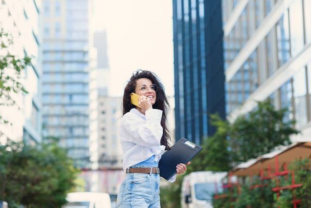 Young business woman walking across the road with tablet among the skyscrapers and speaks on the phone. Working day concept. - 写真・画像