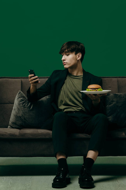 Un joven con un elegante traje verde está comiendo una hamburguesa mientras está sentado en un sofá sobre un fondo verde. Concepto de comida rápida. - Foto, imagen