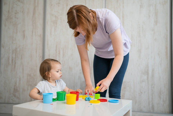 One girl small caucasian toddler child playing with colorful plasticine on the table at home with her mother woman childhood and growing up development concept copy space - 写真・画像