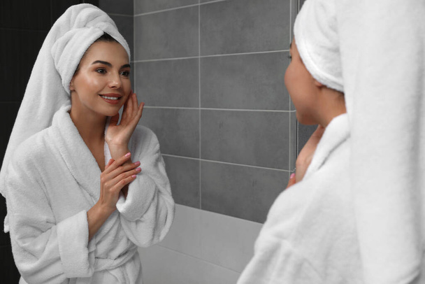 Όμορφη νεαρή γυναίκα σε μπουρνούζι με πετσέτα στο κεφάλι κοντά στον καθρέφτη στο σπίτι - Φωτογραφία, εικόνα