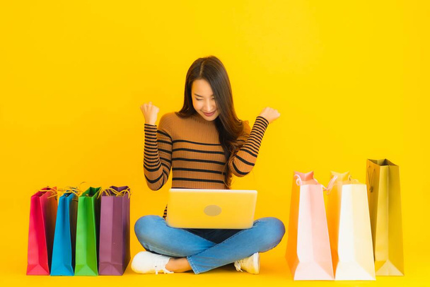 Retrato bonito jovem asiático mulher sentar no chão com laptop ou computador usar cartão de crédito para compras on-line em torno de saco de compras no fundo amarelo
 - Foto, Imagem