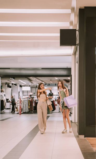 İki güzel arkadaş alışveriş merkezinde yürürken bir tanesi dükkanın camını işaret ediyor. - Fotoğraf, Görsel