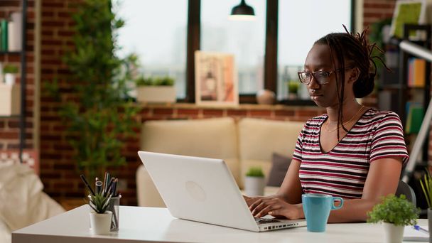 afro-américain freelance dactylographier des informations sur ordinateur portable, travaillant à distance à partir du bureau à domicile. Jeune femme naviguant sur Internet sur ordinateur pour faire du travail à distance, travail d'affaires en ligne à distance. Trépied. - Photo, image