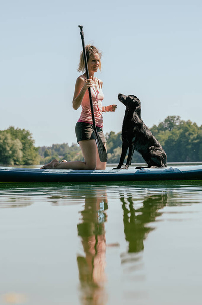 Young female dog owner and her black labrador retriever on a sup board on the river. - Fotó, kép