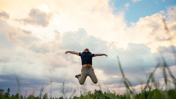 Low angle view through grass blades of a joyful young man jumping high in the air under an evening sky. - Foto, imagen