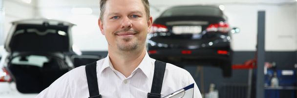 Portrait of qualified maintenance centre worker in special uniform posing on cars background. Mechanic male propose high service. Pit stop, garage concept - Photo, Image