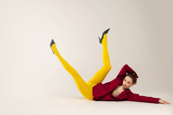 Portret van jong stijlvol meisje in gele maillot, rode jas en hiel schoenen poseren op de vloer geïsoleerd over grijze studio achtergrond. Concept van retro mode, kunst fotografie, stijl, queer, schoonheid - Foto, afbeelding