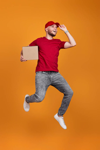 Delivery man in red cap, t-shirt holding cardboard box and jumping isolated on orange background - Foto, immagini