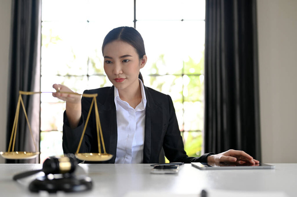 Schöne und professionelle asiatische Anwältin oder Rechtsberaterin sitzt an ihrem Bürotisch, hält eine Waage der Gerechtigkeit in der Hand. Recht und Rechtskonzept - Foto, Bild