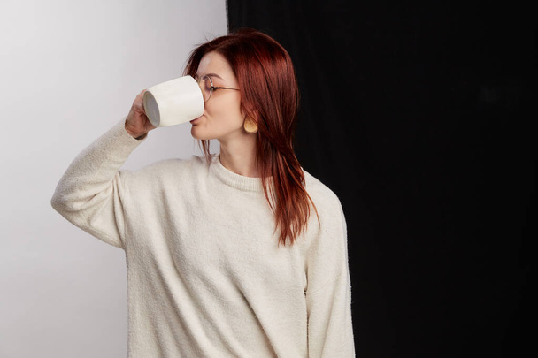 A hot drink invigorates. Redhead woman holding cup of tea or coffee in hand, face expression stands on half white and black background in big eyeglasses - Foto, Imagen